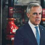 Prime Minister Mark Carney in a dark suit and blue tie, photographed in front of industrial pipefitting equipment at a Calgary union training facility.
