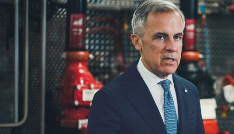 Prime Minister Mark Carney in a dark suit and blue tie, photographed in front of industrial pipefitting equipment at a Calgary union training facility.